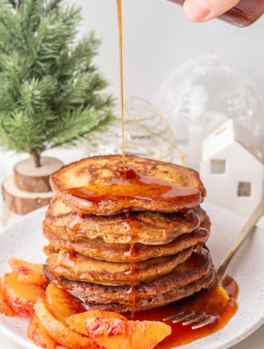 Orange gingerbread syrup is being poured over a stack of gingerbread pancakes on a white plate.