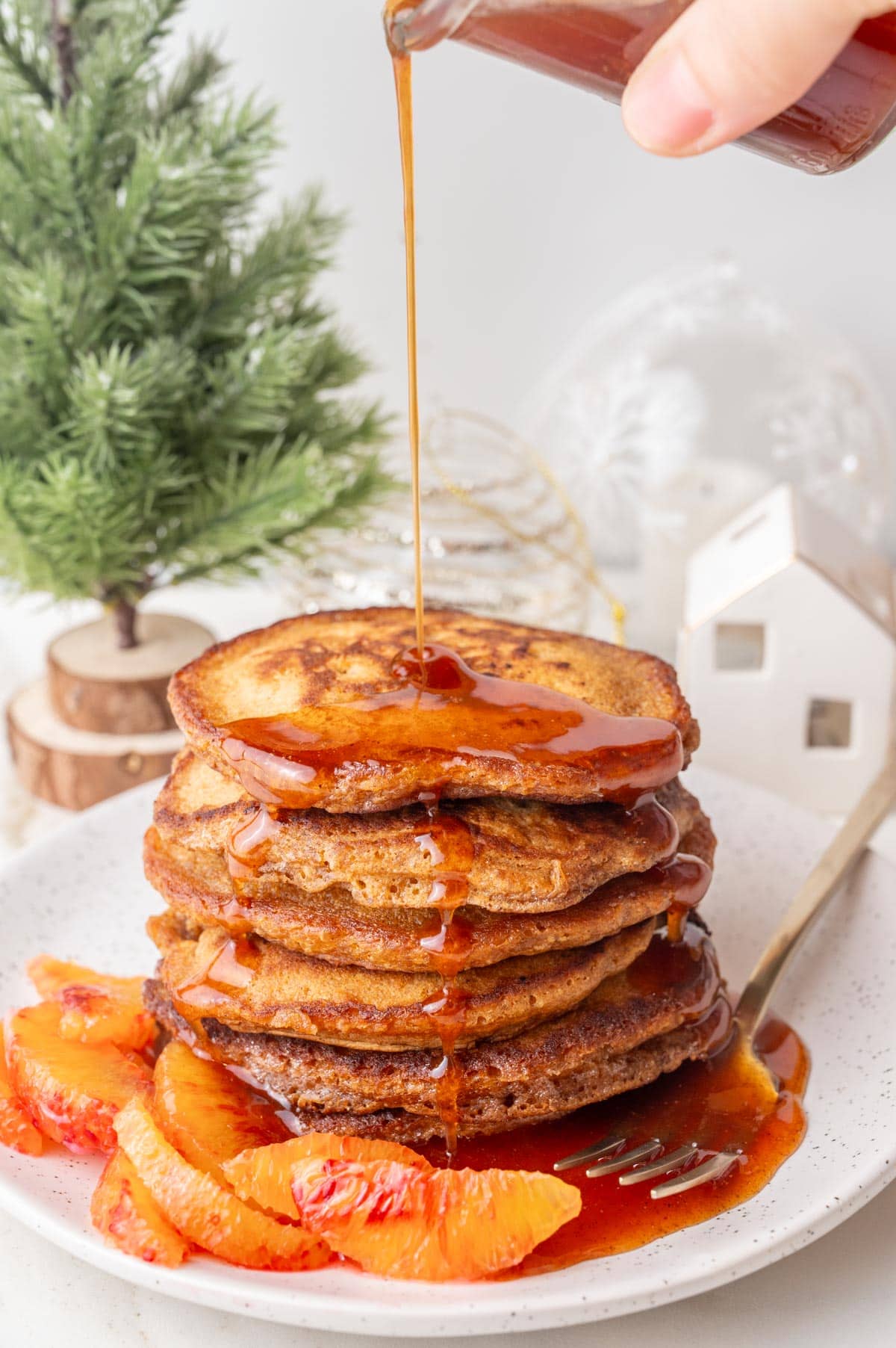 Orange gingerbread syrup is being poured over a stack of gingerbread pancakes on a white plate.