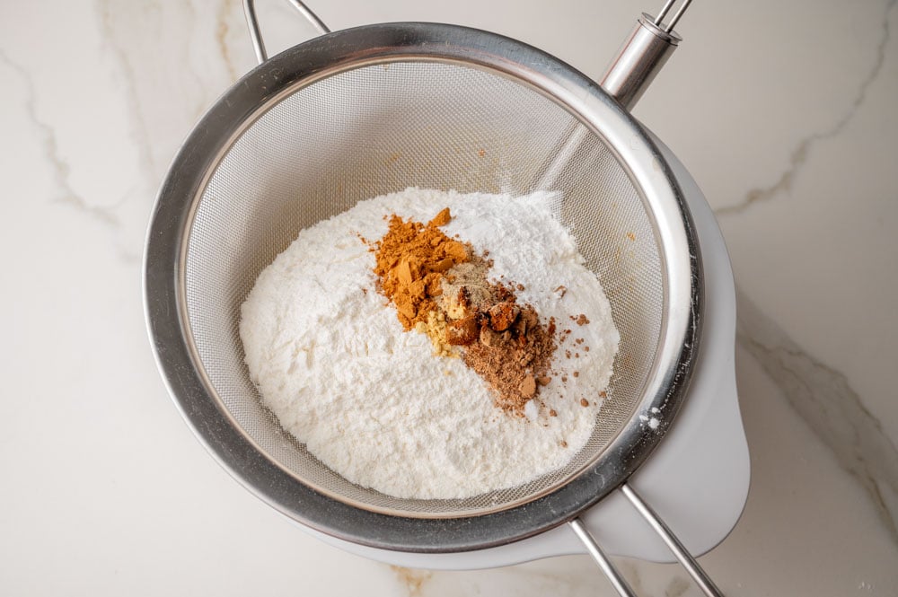 A sieve with flour and spices hung over a white bowl.