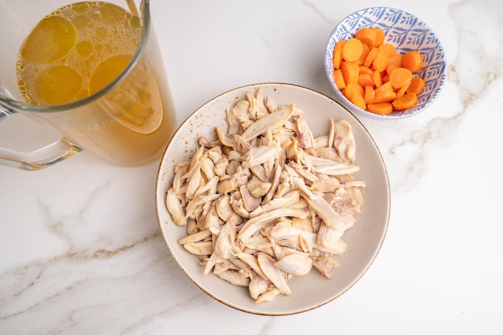 Shredded cooked chicken in a bowl, chicken broth in a pitcher, and cooked carrots in a bowl.