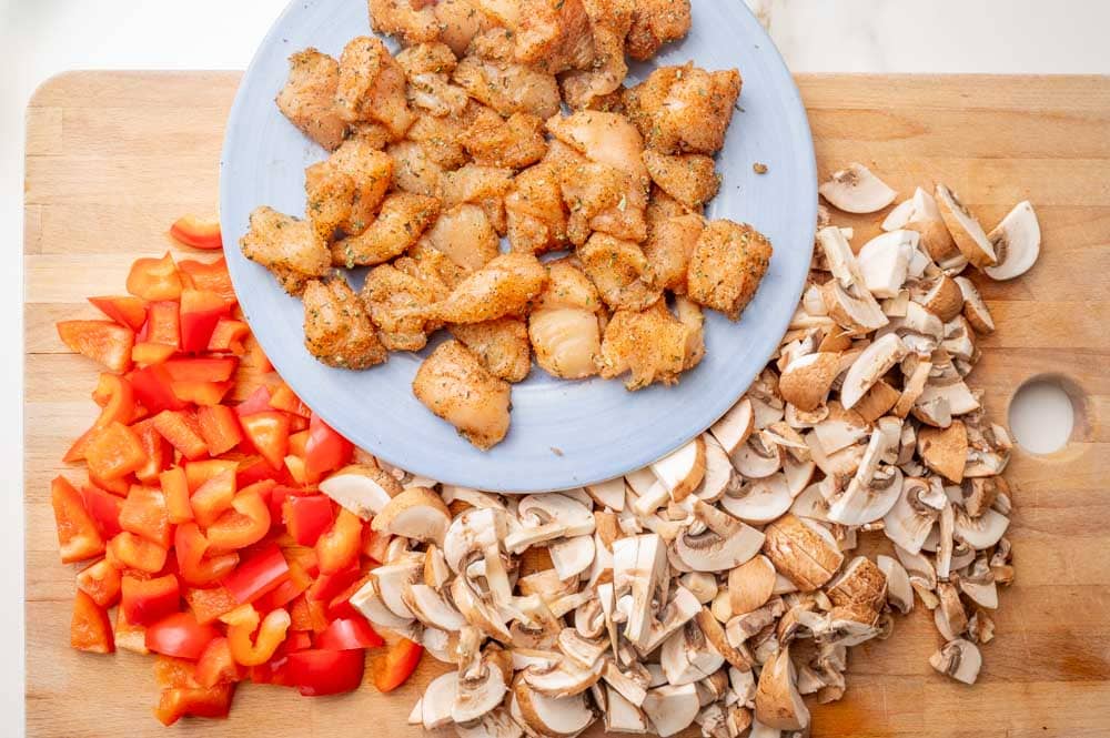Chopped bell pepper and mushrooms on a wooden board. Diced seasoned chicken on a plate.
