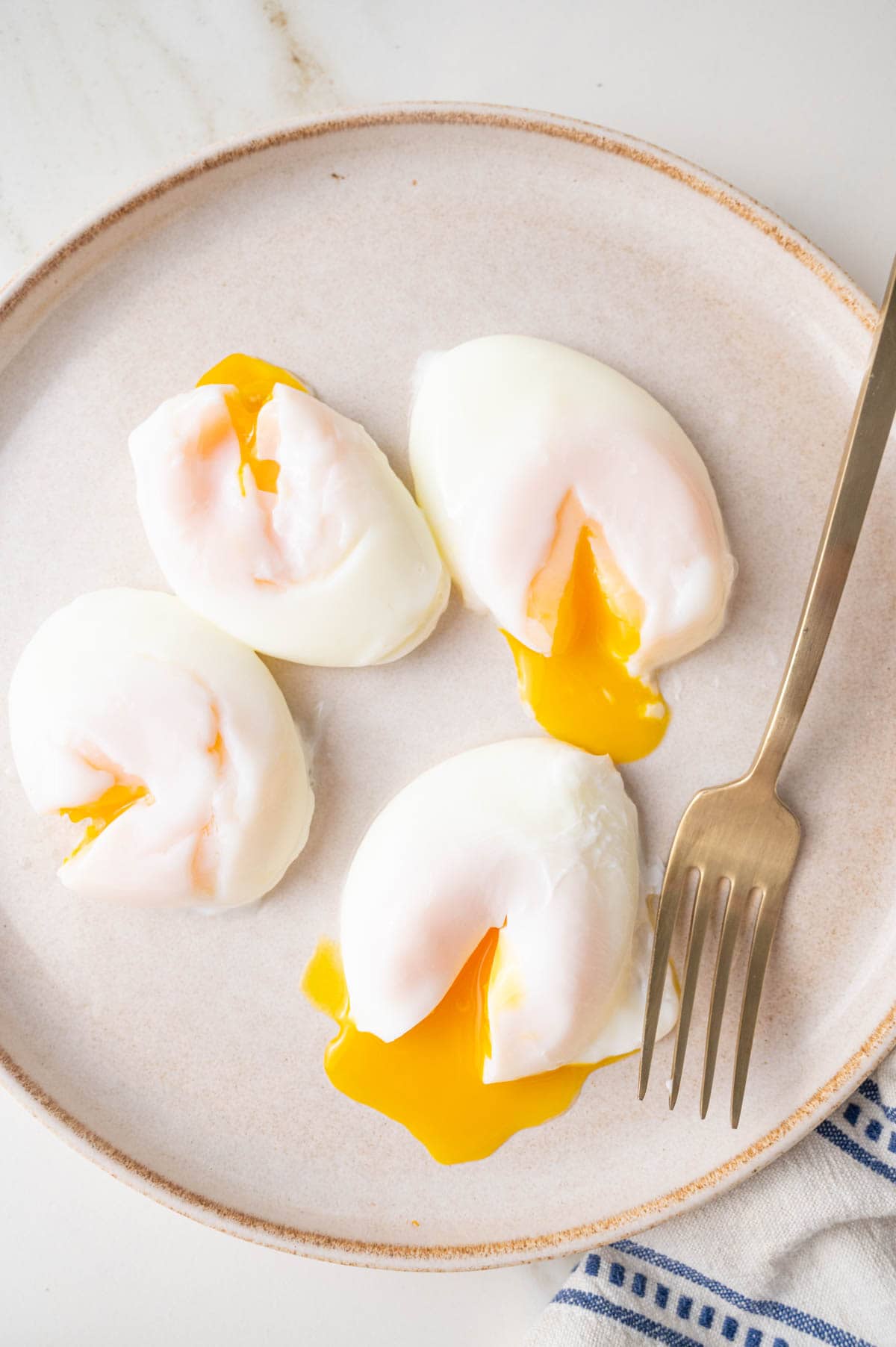 Four poached eggs with runny egg yolks on a beige plate. Gold fork on the side.