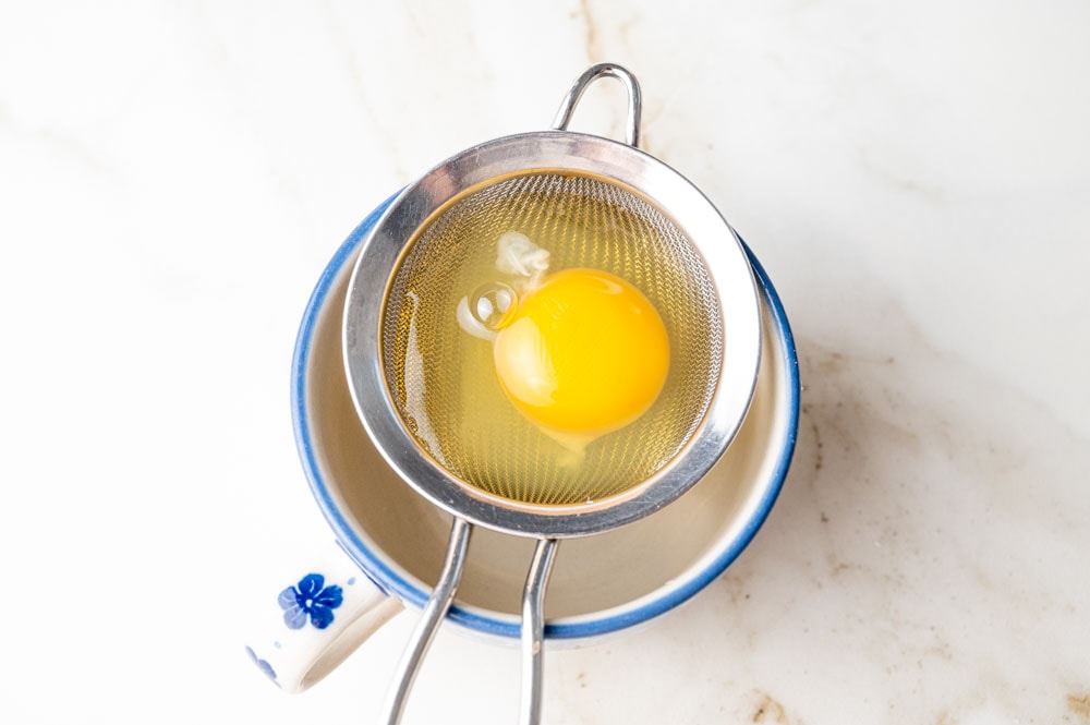 A raw egg placed atop a small fine-mesh strainer suspended over a small cup.