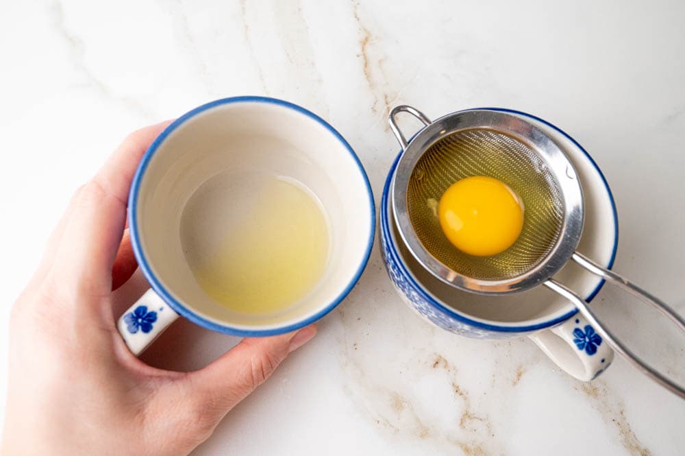 A raw egg placed atop a small fine-mesh strainer suspended over a small cup and a second small cup with the drained liquid from the egg white.