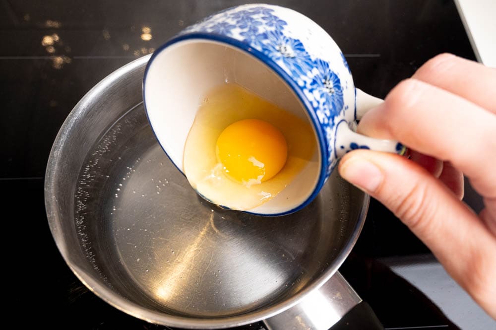 A raw egg is being added to a pot with simmering water.