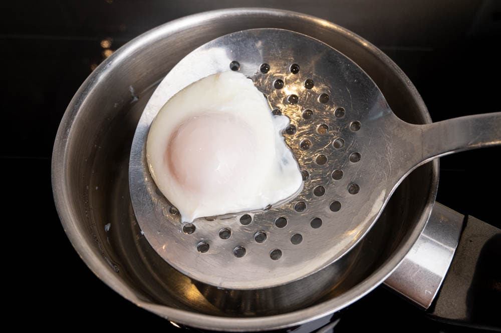 A poached egg is being lifted from the water with a slotted spoon.