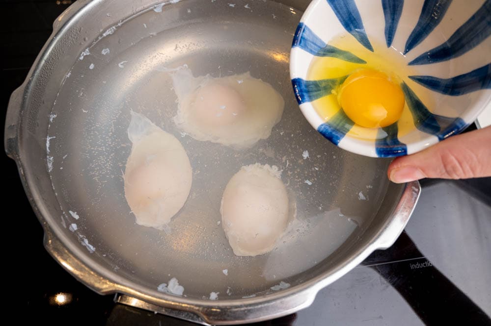 A raw egg is being added to a large pot with simmering water and 3 eggs.