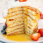 A stack of pancakes on a white plate. Maple syrup is being poured over the pancakes. Berries on the side.