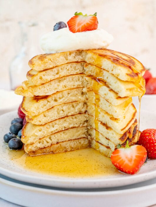 A stack of pancakes on a white plate. Maple syrup is being poured over the pancakes. Berries on the side.