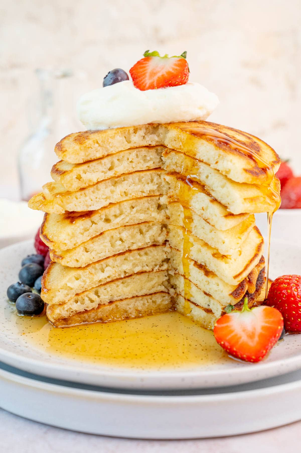 A stack of pancakes with a part missing on a white plate. Maple syrup is being poured over the pancakes. Berries on the side.