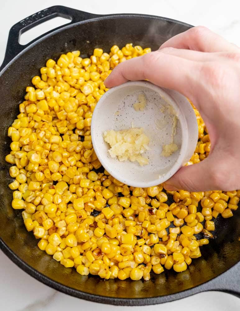 Pan-fried corn kernels in a cast iron pan are being seasoned with garlic.