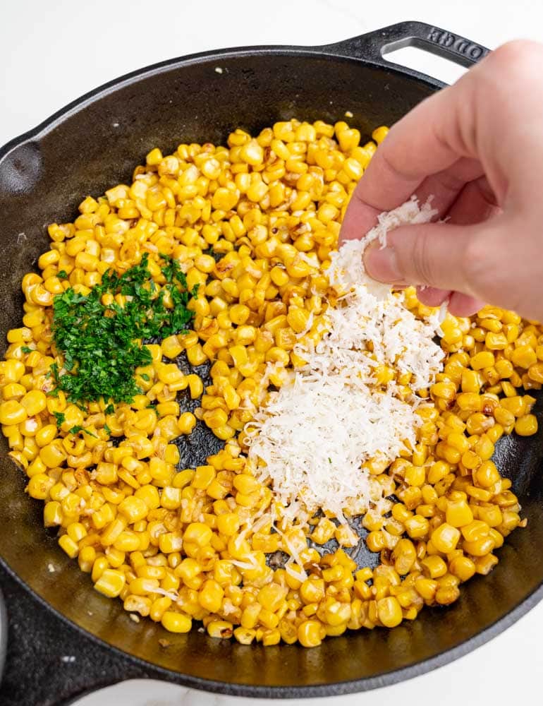 Pan-fried corn kernels in a cast iron pan are being seasoned with parley and parmesan.