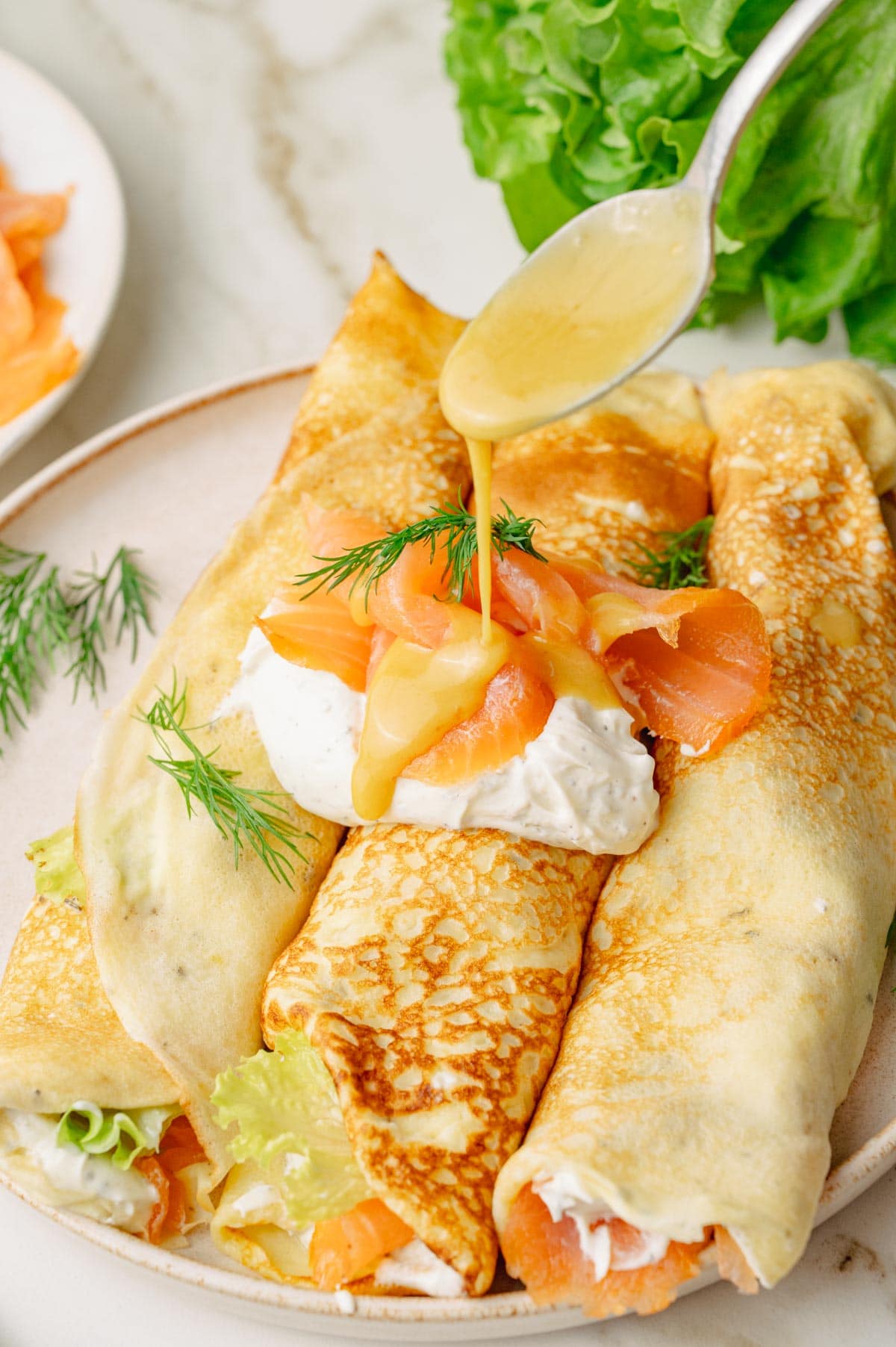 Honey mustard sauce is being poured over smoked salmon crepes on a beige plate.