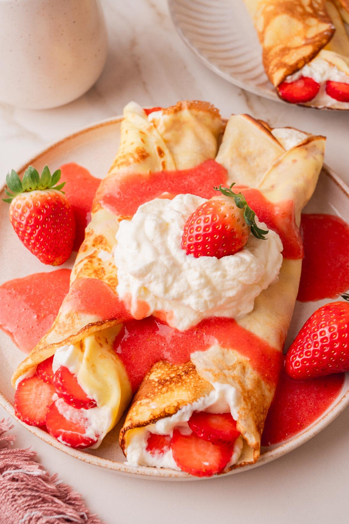 Crepes filled with whipped cream and fresh strawberries served with strawberry sauce on a beige plate.