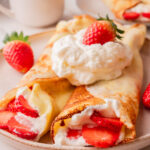 Crepes filled with whipped cream and strawberries served with strawberry sauce on a beige plate.