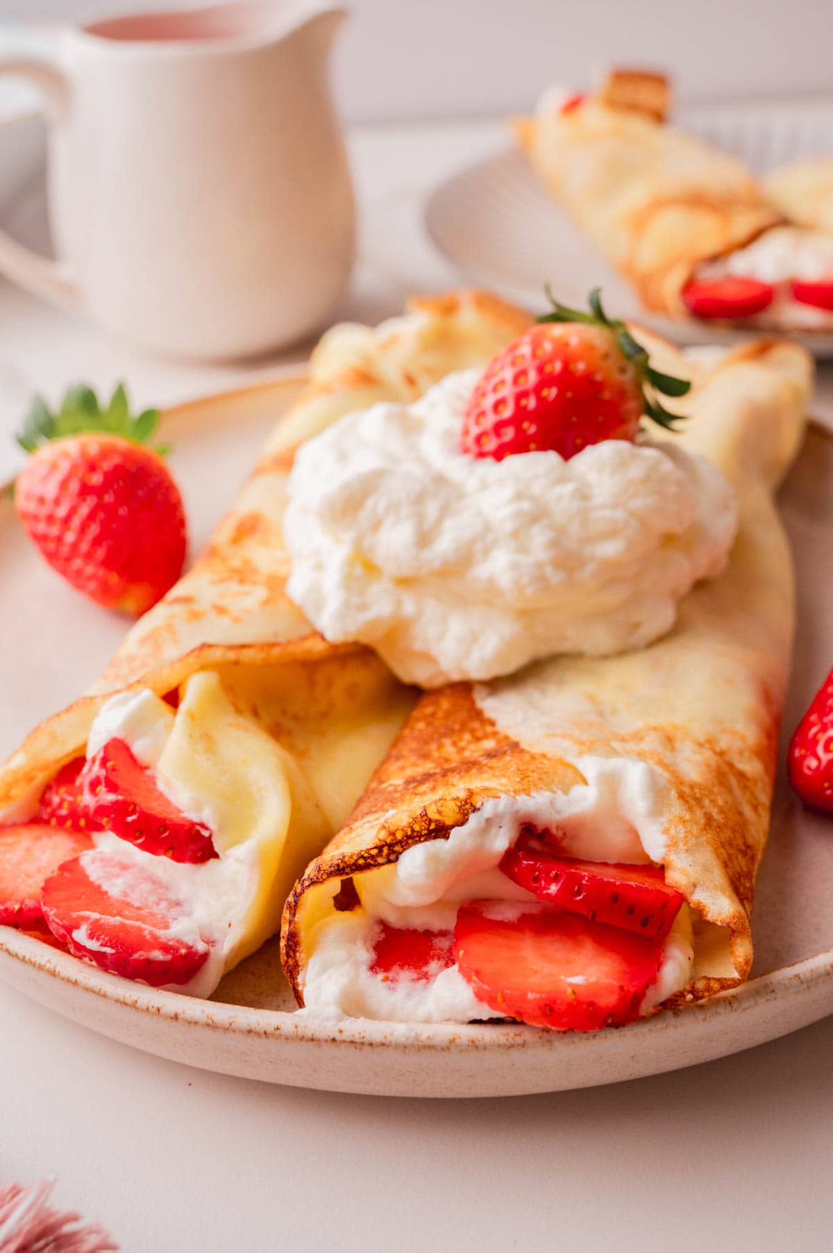 A close up photo of crepes filled with whipped cream and fresh strawberries served with strawberry sauce on a beige plate.