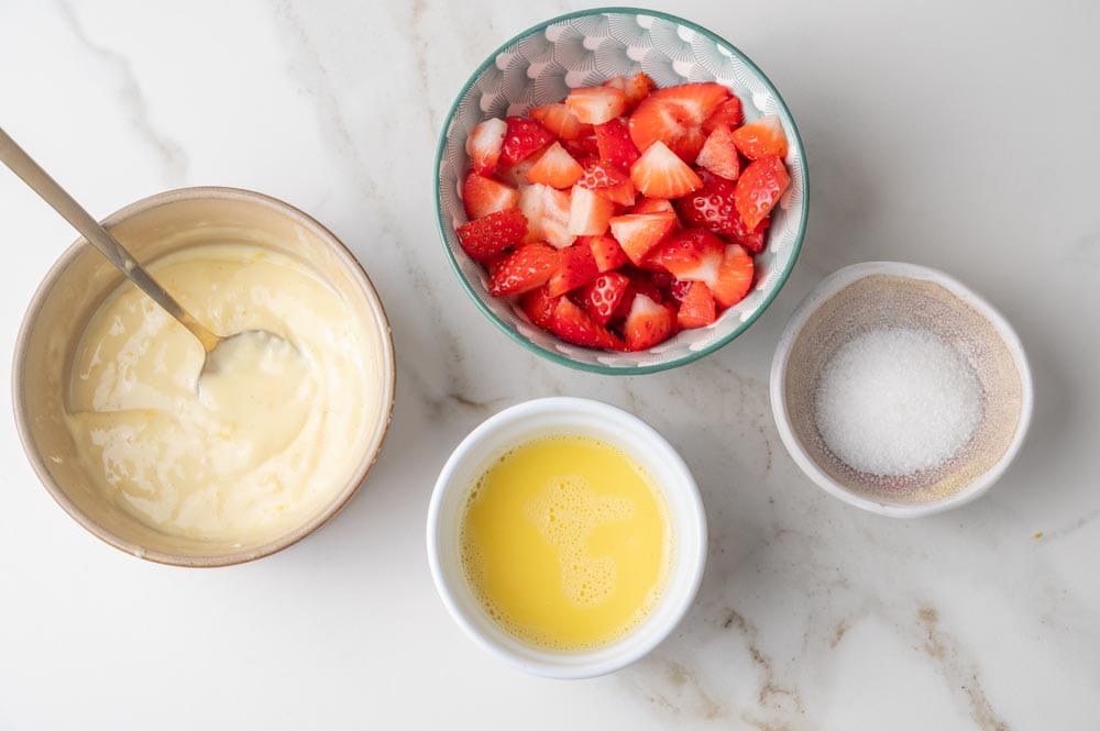 Cream cheese filling in a bowl, chopped strawberries in a bowl, egg wash in a bowl, granulated sugar in a bowl.