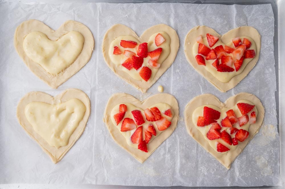 Heart-shaped puff pastry on a parchment paper topped with cream cheese layer and strawberries.