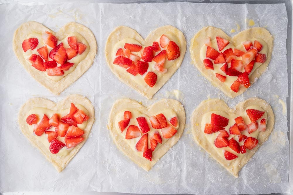 Heart-shaped puff pastry on a parchment paper topped with cream cheese layer and strawberries.