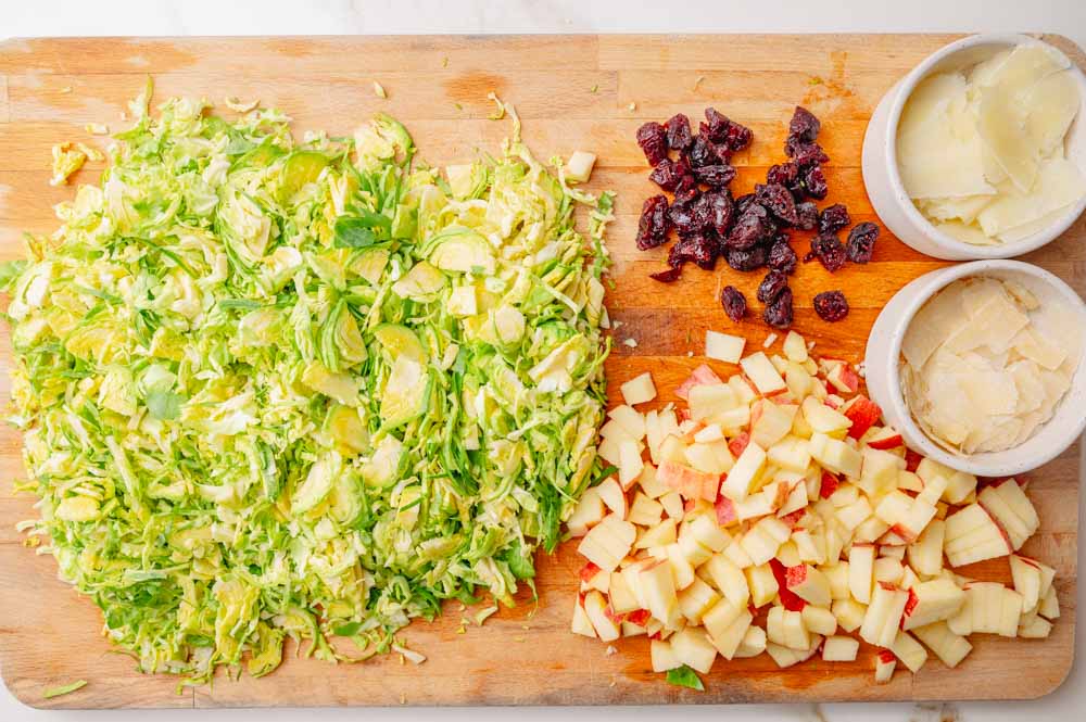 Shaved brussel sprouts, shaved parmesan and pecorino, chopped apples and cranberries on a wooden chopping board.