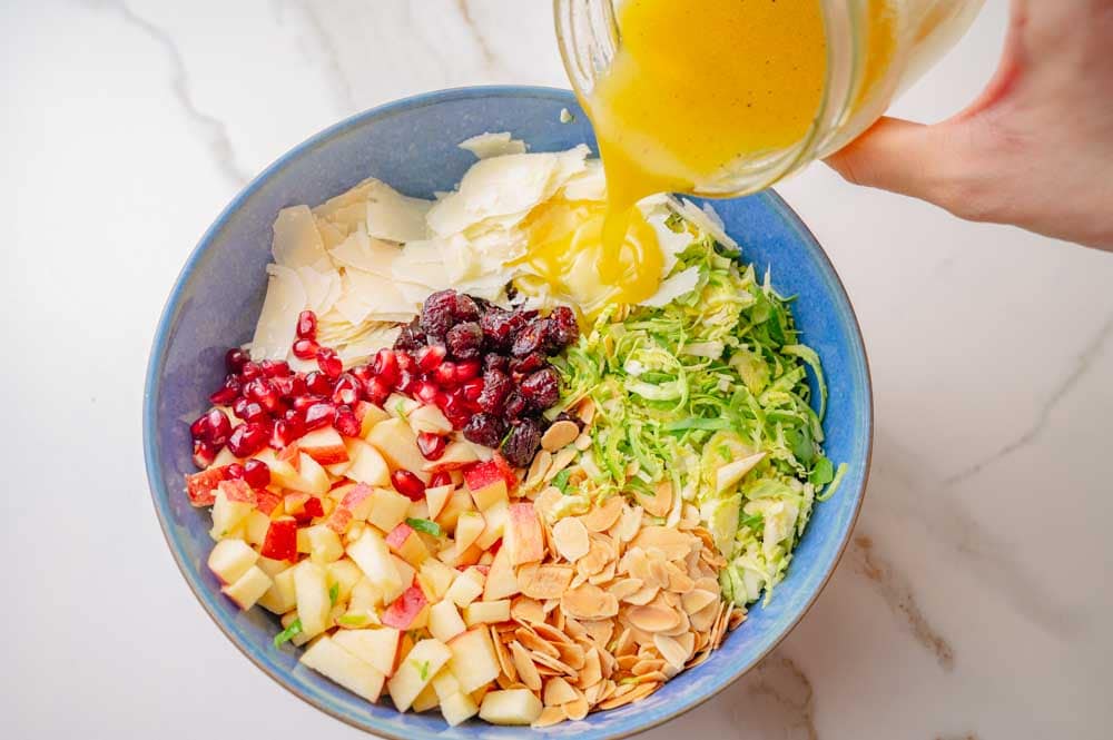 Salad dressing is being added to brussel sprouts salad ingredients in a blue bowl.