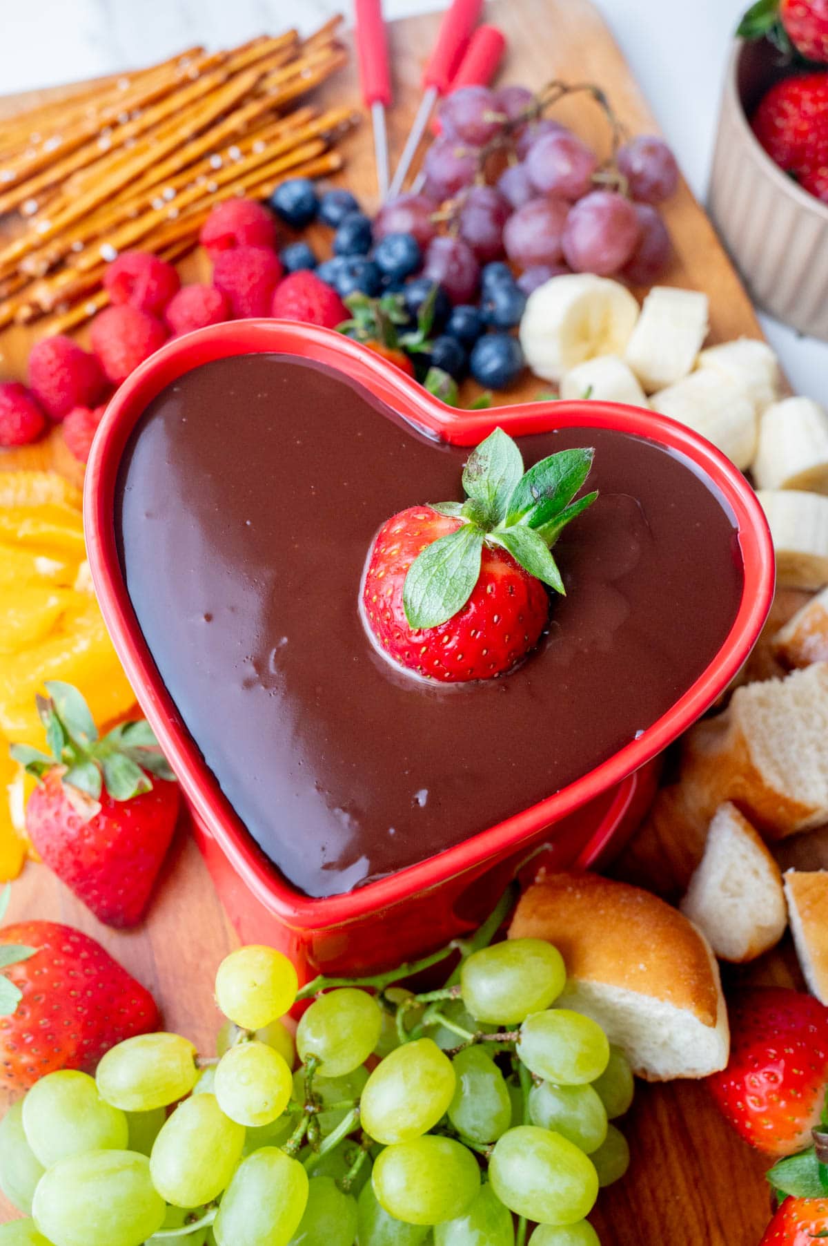 Chocolate fondue in a heart-shaped fondue bowl surrounded with fruit and baked good on a wooden board.