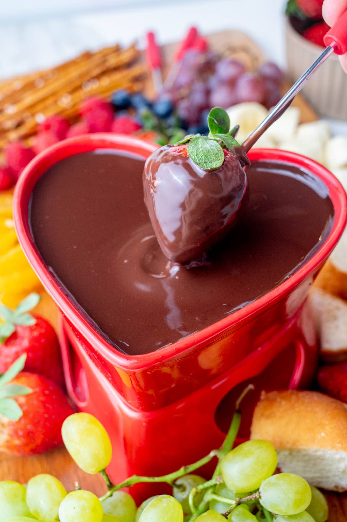 A strawberry dipped in chocolate fondue in a heart-shaped fondue bowl surrounded with fruit and baked good on a wooden board.