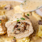 Cut in half mushroom-stuffed chicken breast is being poured with creamy sauce.