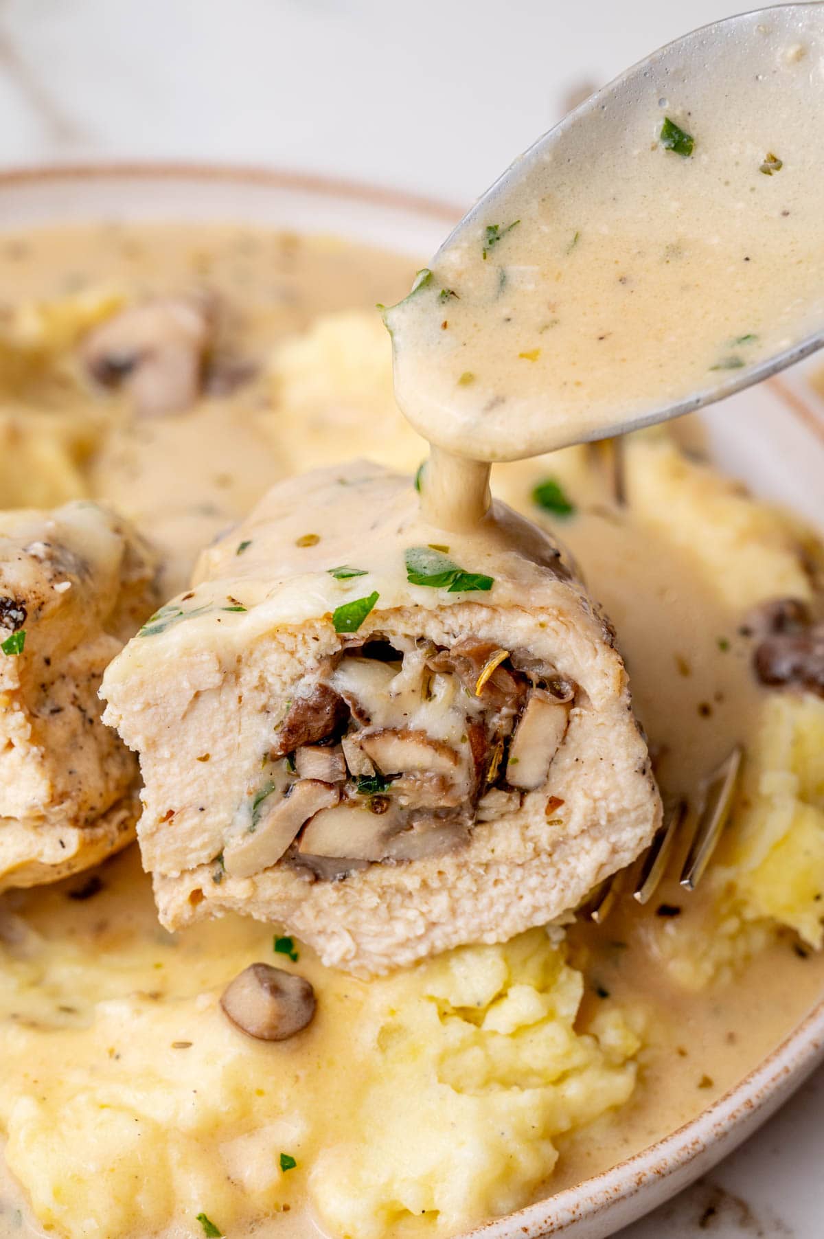 Cut in half mushroom-stuffed chicken breast and mashed potatoes are being poured with a creamy sauce.