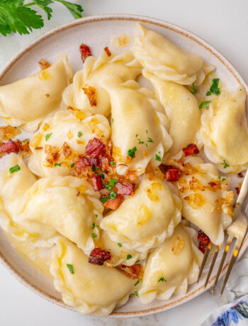 Pierogi ruskie topped with bacon and onions on a beige plate.