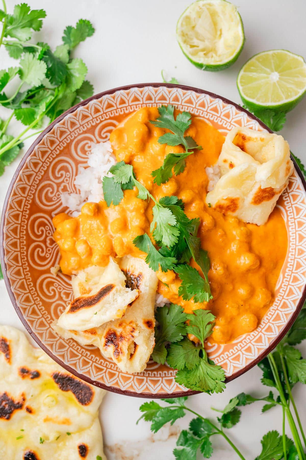 Chickpea curry with rice, Naan bread pieces, and cilantro in a brown bowl. Limes and cilantro leaves around the bowl.