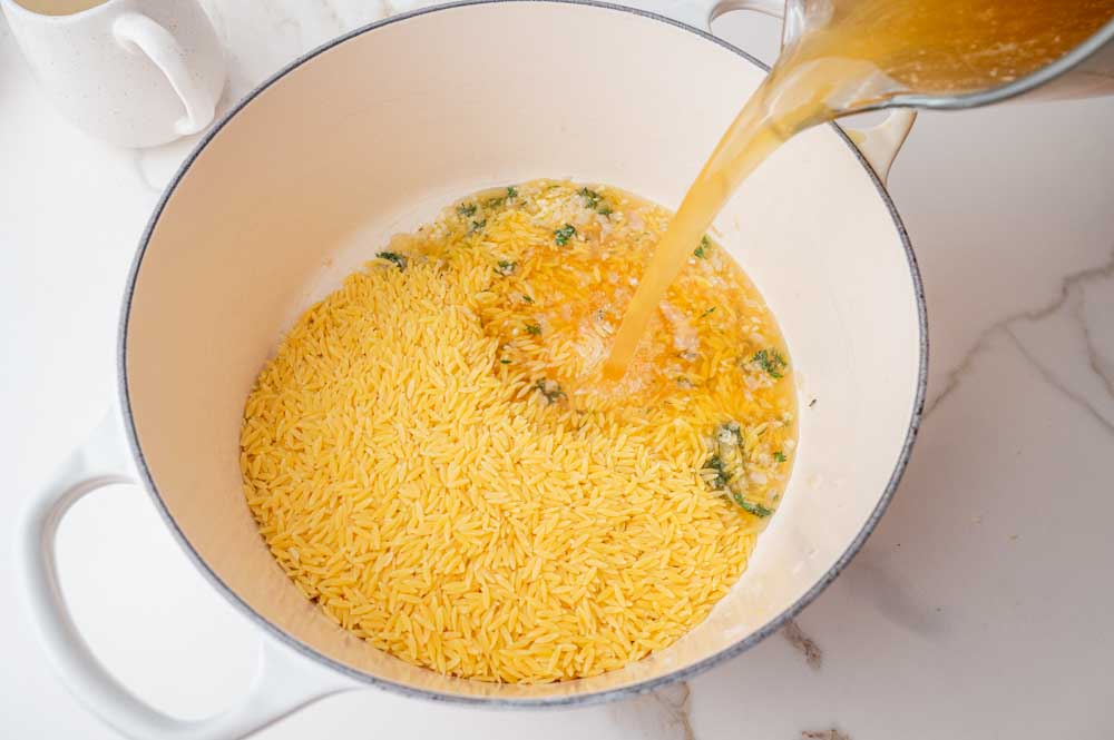 Broth is being added to orzo in a white pot.