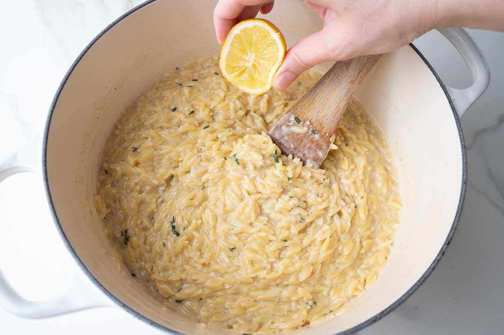 Creamy orzo in a white pot is being seasoned with lemon juice.