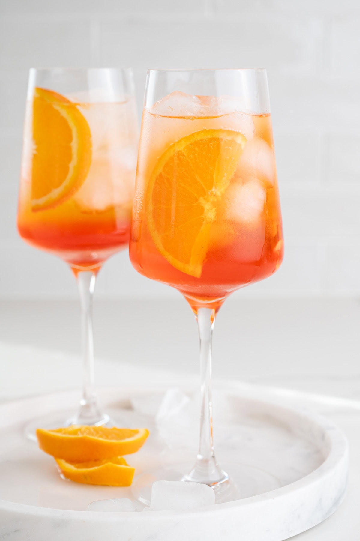 Two glasses with Aperol Spritz cocktail garnished with orange slices on a white marble board.