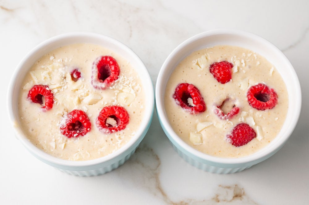 Baked oats batter with white chocolate chunks and raspberries in two ramekins.