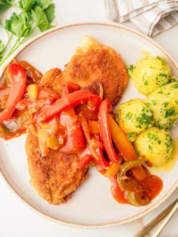 Zigeunerschnitzel with bell pepper sauce and parsley potatoes on a beige plate.