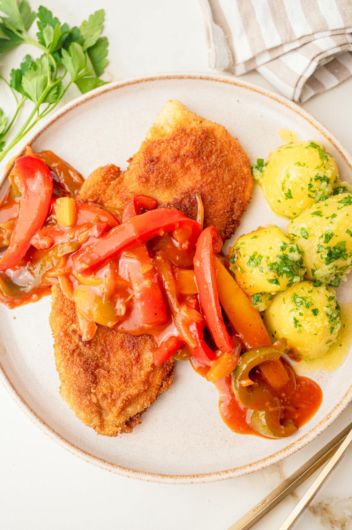 An overhead photo of breaded chicken cutlets with bell pepper and onion sauce and parsley potatoes on a plate.