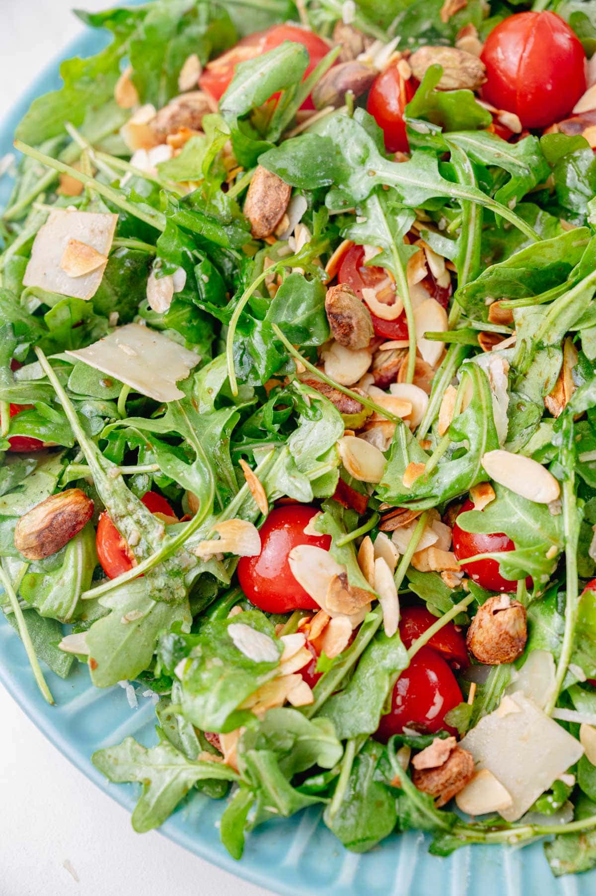 A close up photo of arugula salad with tomatoes, nuts, and parmesan on a blue plate.
