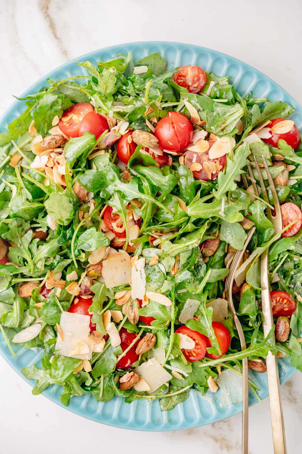 An overhead photo of arugula salad with tomatoes, nuts, and parmesan on a blue plate.