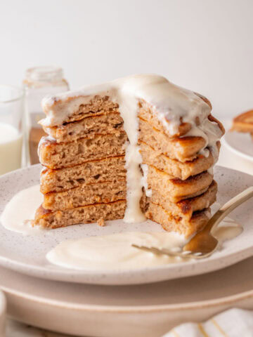 A tall stack of fluffy cinnamon pancakes with a generous cream cheese glaze dripping down the sides, served on a white speckled plate with a fork and extra glaze on the side. A large wedge is cut out, showing the soft, airy texture inside.