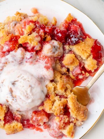 Close-up of a serving of strawberry rhubarb crumble on a plate with melted ice cream and a spoon.
