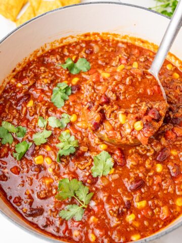 Hearty chili con carne with ground beef, beans, corn, and tomatoes simmering in a white Dutch oven, garnished with fresh cilantro and served with tortilla chips.