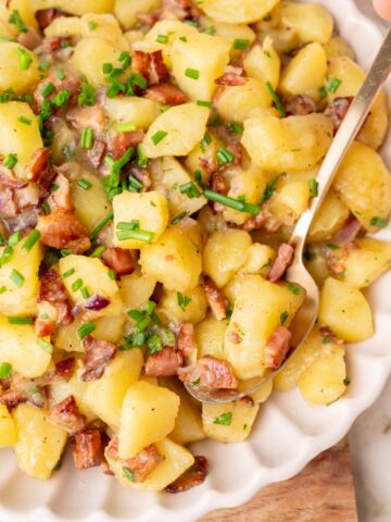 German potato salad topped with bacon and chives in a beige bowl.